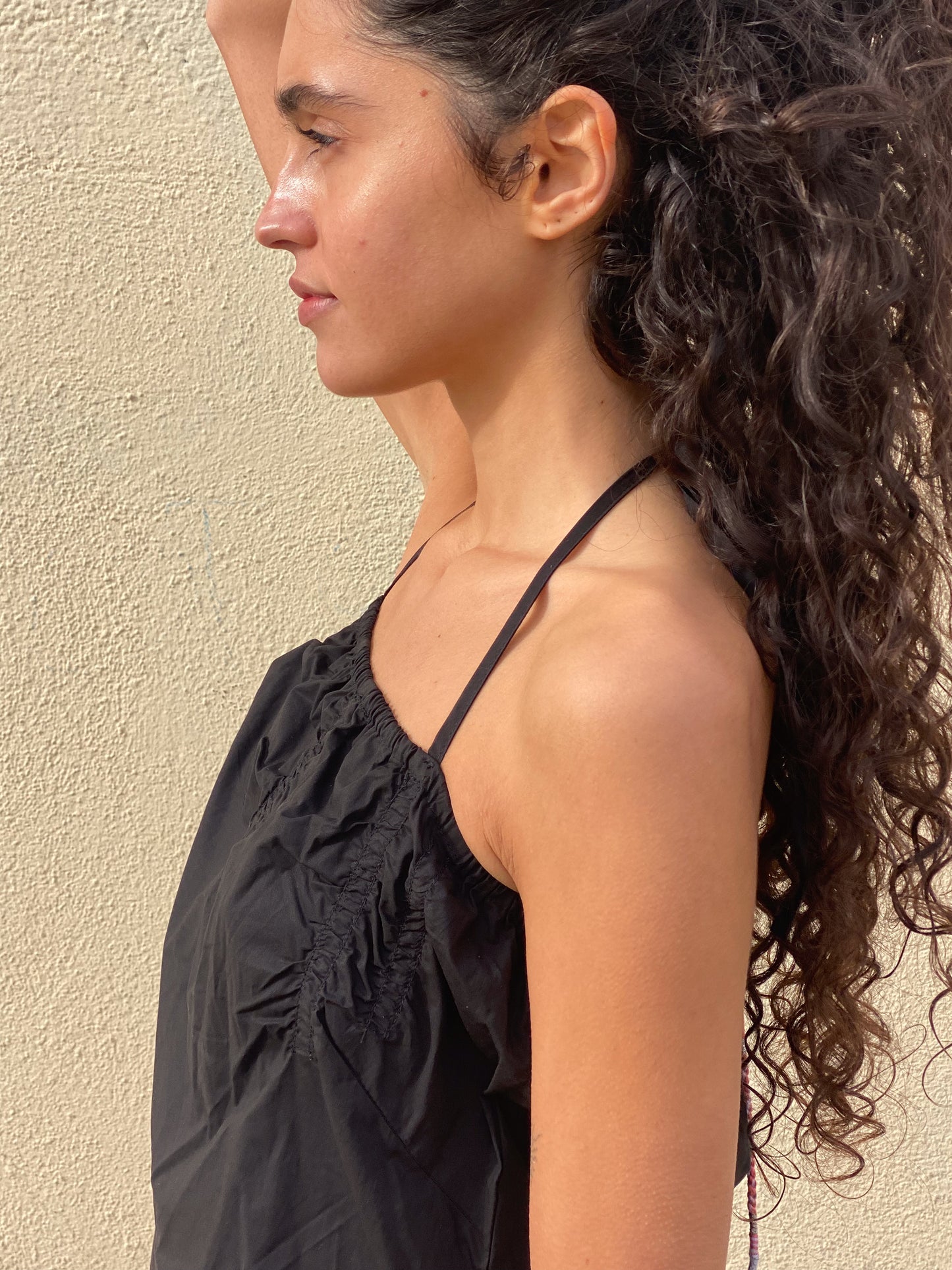 A woman with long, curly hair stands in profile against a textured beige wall, wearing the sleek Tube Top - Black by Deiji Studios. One arm is raised above her head as she gazes forward with a calm expression.