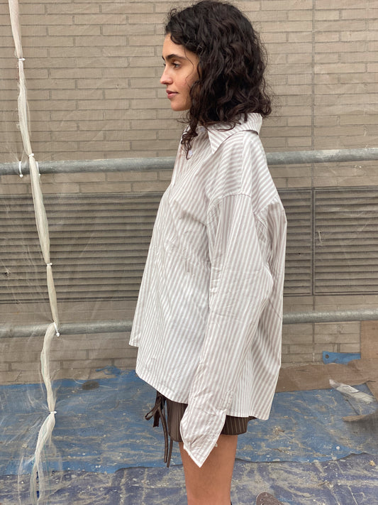 A person with curly dark hair stands in profile against a brick wall, wearing an oversized, light-colored striped day shirt and brown shorts. Blue material covers the ground, with metal rails and plastic sheets behind them.