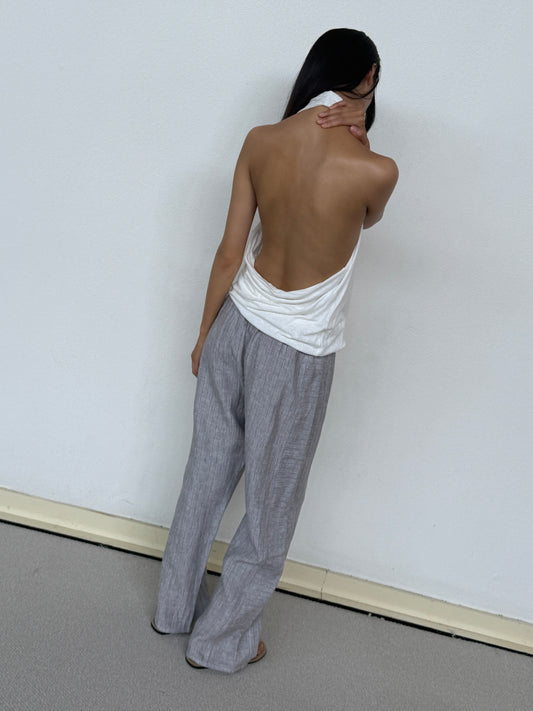 A person stands barefoot facing a white wall, wearing 0s Double Waist Linen Pant in Haze Blue and a backless white top, with one hand touching the back of their neck.