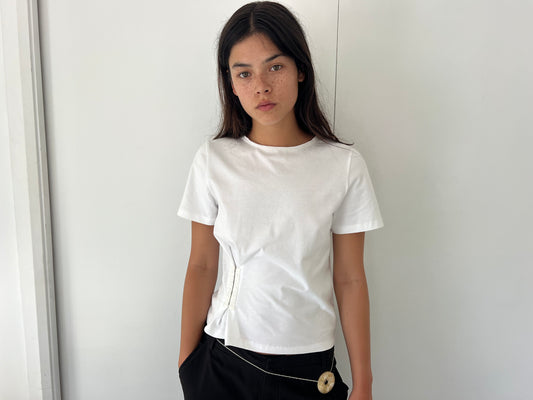 A young woman with long dark hair and freckles stands against a white wall, wearing the Pinch Tee - White by Deiji Studios and black pants, one hand in her pocket and a neutral expression.