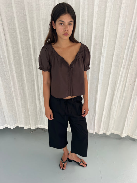 A young woman with long dark hair stands indoors on a light grey floor before white curtains, wearing a Deiji Studios Close Shirt in Sepia, black wide-leg pants, and black sandals. She gazes slightly to the side with a neutral expression.