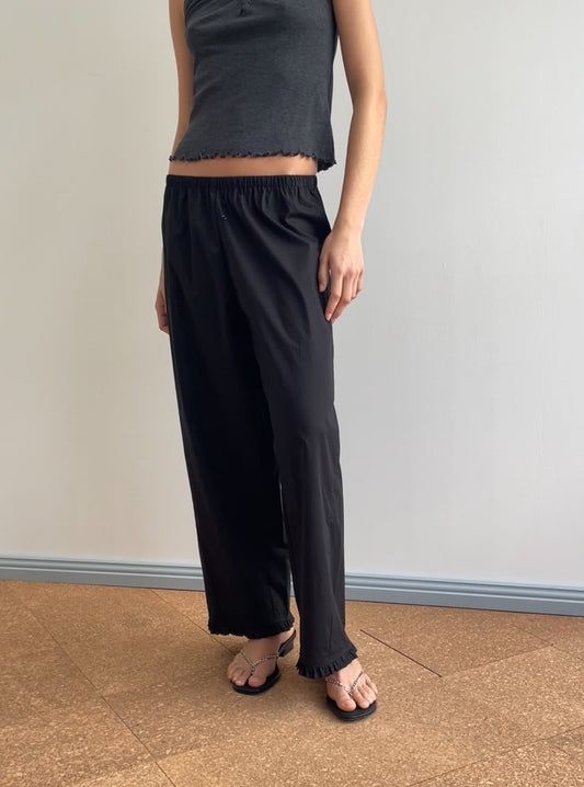 A person stands indoors on a cork floor by a light wall, wearing a sleeveless dark gray crop top, Deiji Studios Flounce Pant - Black with an elastic waistband, and black sandals.