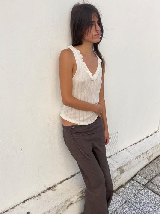 A young woman with long brown hair stands against a white wall, wearing a sleeveless white top with ruffled edges and the Deiji Studios Double Belted Pant - Brown Stripe. She gazes off to the side with a neutral expression.