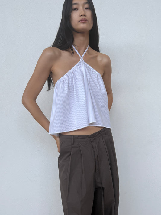 A person with long dark hair wears a sleeveless halter-neck white top and the Turn Pant in Cocoa from Deiji Studios, standing against a plain light-colored background.