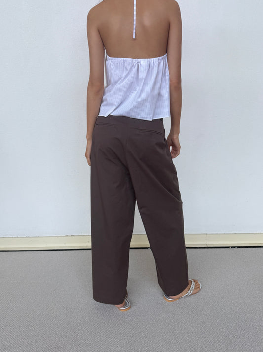 A person stands with their back to the camera, barefoot on a light carpet against a white wall, wearing Deiji Studios’ Turn Pant in Cocoa—relaxed and tailored with an adjustable drawcord—and a backless, halter-style white top.