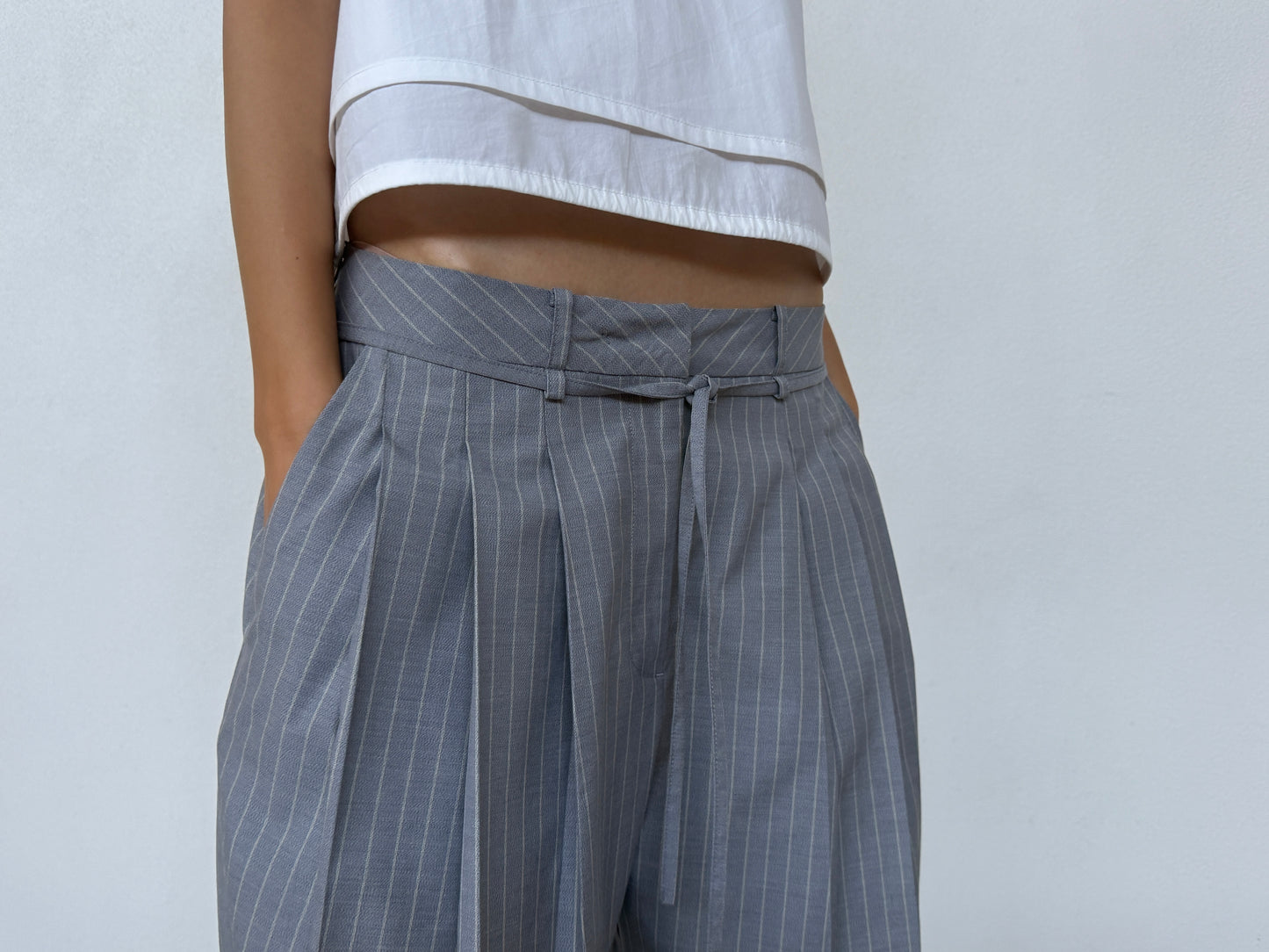 A person wearing Deiji Studios Tri Pleat Pure Wool Pant - Steele Stripe with hands in pockets and a white top, standing against a plain light background.