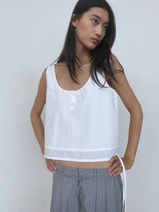 A woman with long dark hair wears the Layered Top - White, a sleeveless organic cotton piece with buttons, paired with light gray pinstriped pants. She stands against a plain white wall, looking to the side with a relaxed expression.