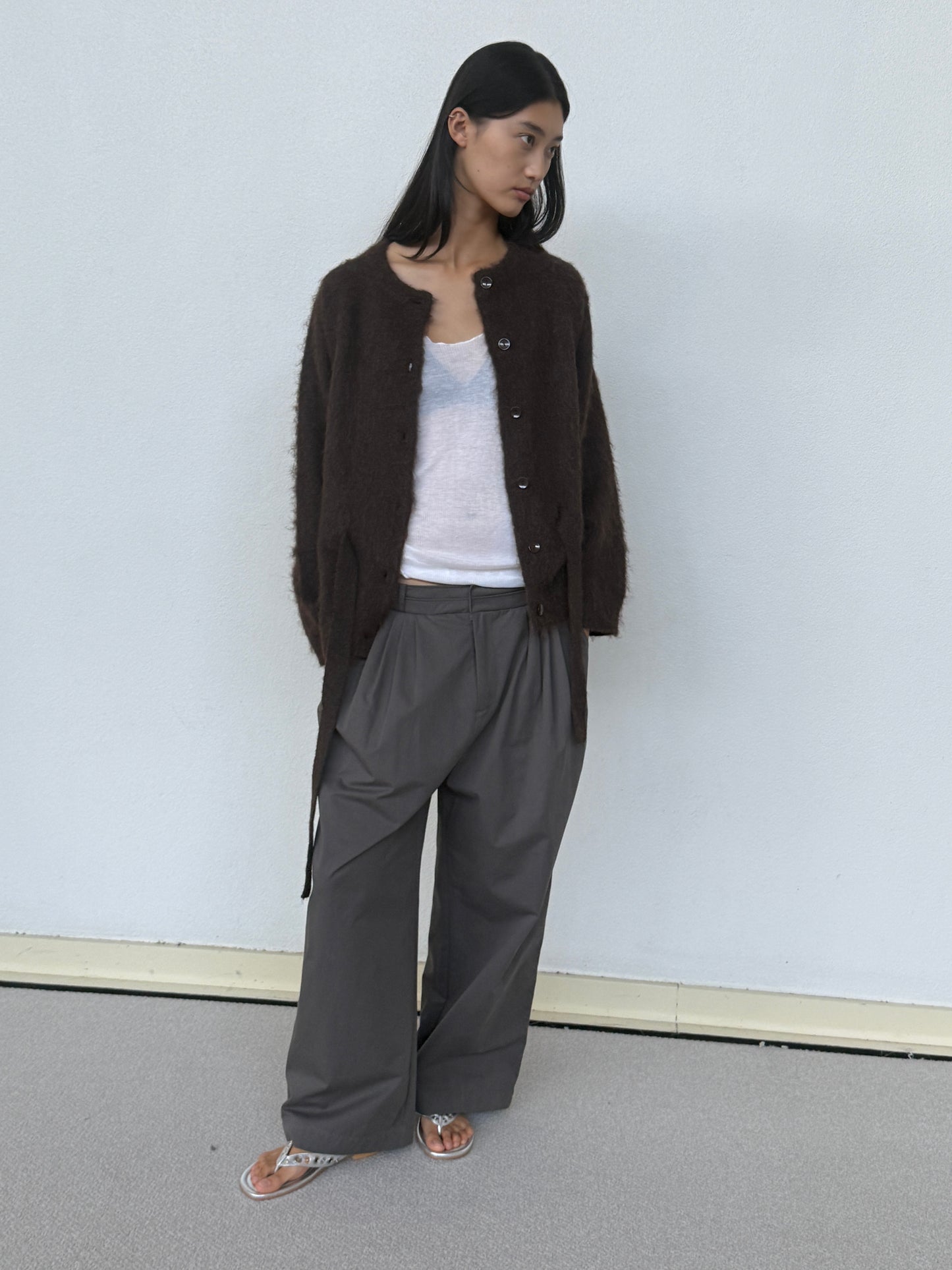 A woman stands against a light wall, wearing a dark brown fuzzy cardigan over a white top and 0’s Two Layer Pant in Charcoal. She completes the look with silver sandals, her long dark hair flowing as she looks left.