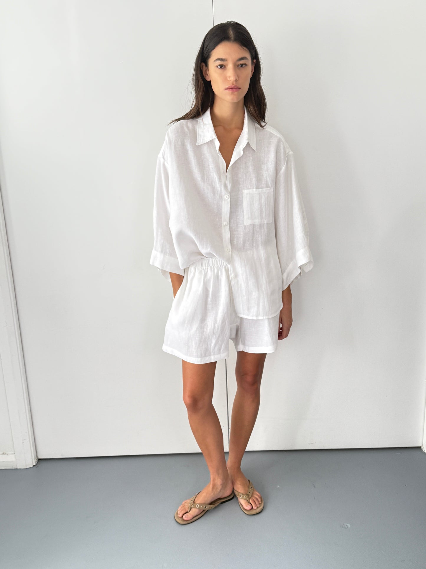 A woman with long dark hair and a neutral expression stands indoors against a white wall, wearing Deiji Studios the 03 set in white—an organic linen shirt with rolled sleeves and matching boxer-style shorts—paired with tan sandals.