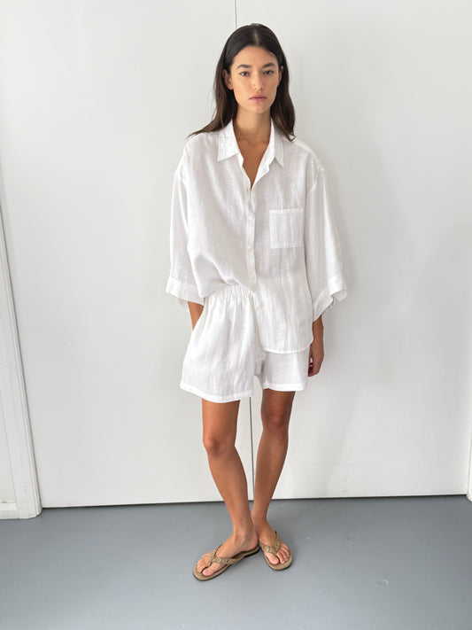 A woman with long dark hair and a neutral expression stands against a white wall, wearing the 03 set - white by Deiji Studios, featuring a loose linen shirt with rolled sleeves and matching boxer shorts, paired with tan sandals.