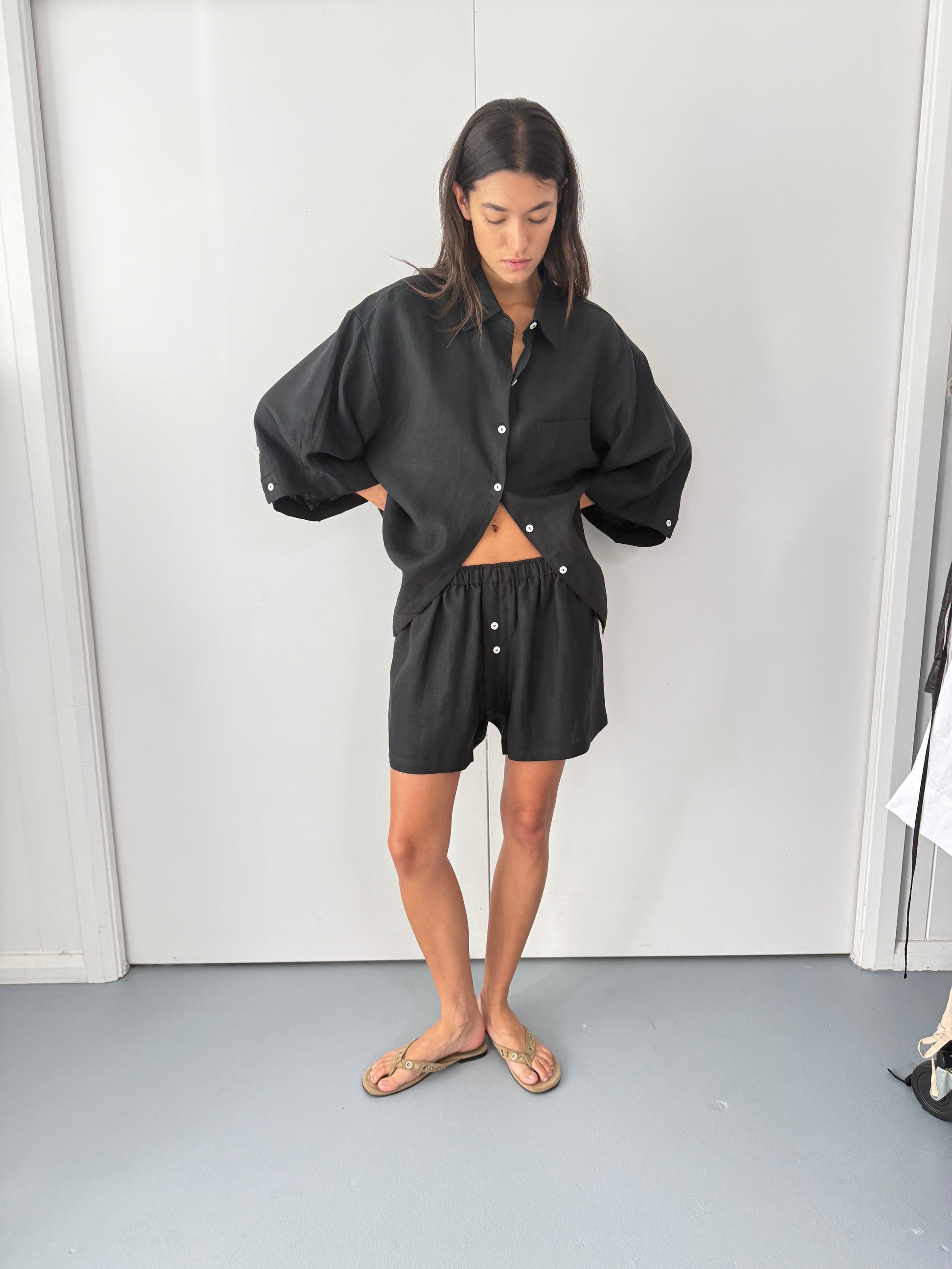 A person stands indoors wearing Deiji Studios the 03 set - deep black, an oversized linen shirt with boxer-style shorts, paired with beige sandals and hands in pockets, against a plain white background.