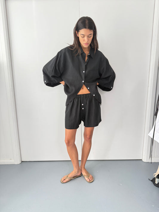 A person stands indoors wearing Deiji Studios the 03 set - deep black, an oversized linen shirt with boxer-style shorts, paired with beige sandals and hands in pockets, against a plain white background.