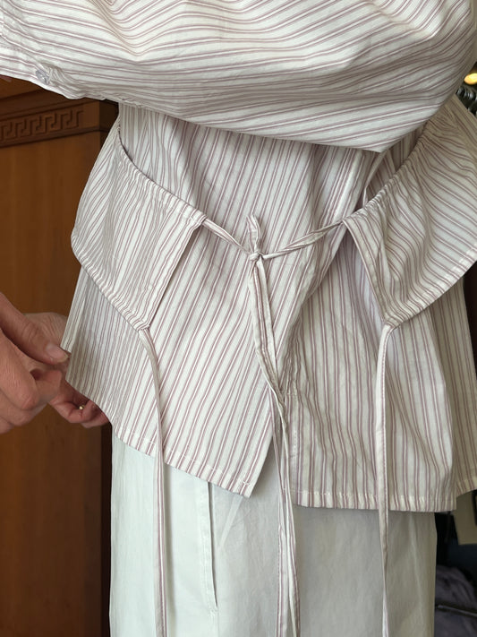 A close-up shows a person wearing the Cotton Sash, a light-striped, long-sleeved shirt with waist ties. Another hand adjusts it on the left. White pants and part of a wooden cabinet are also visible.