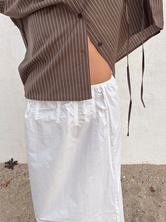 A person in a brown pinstriped, partially unbuttoned shirt and the Channel Skirt stands on sand against a textured off-white wall, showcasing part of their waist and hip.