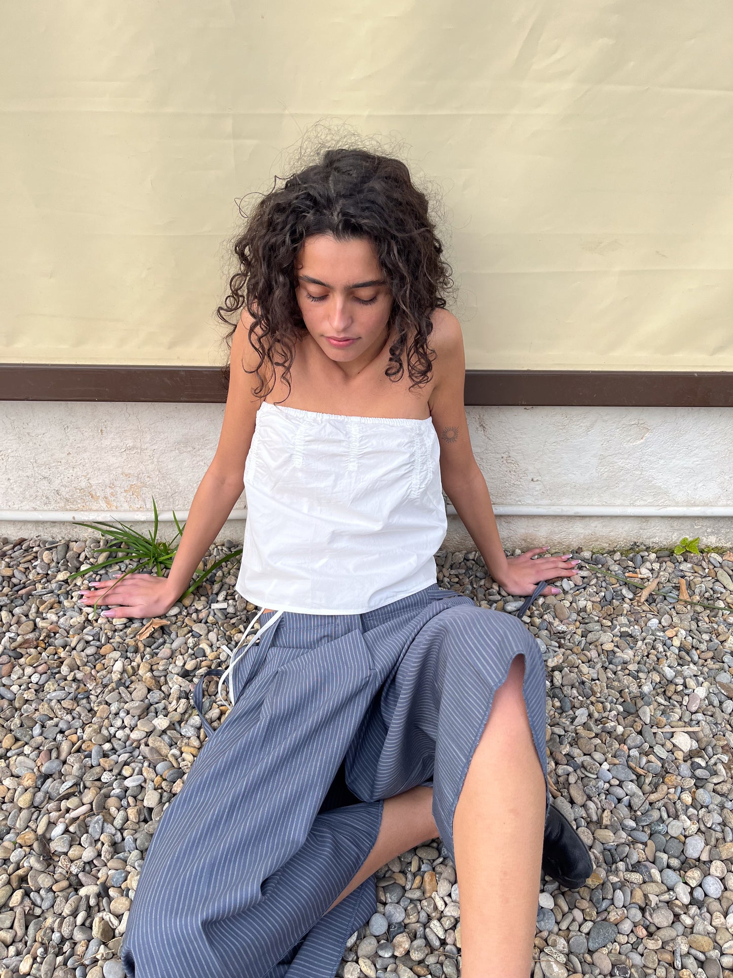 A woman with curly hair sits on gravel by a beige wall, wearing a Deiji Studios Tube Top - White and wide-legged pinstriped pants, looking down with a relaxed expression.