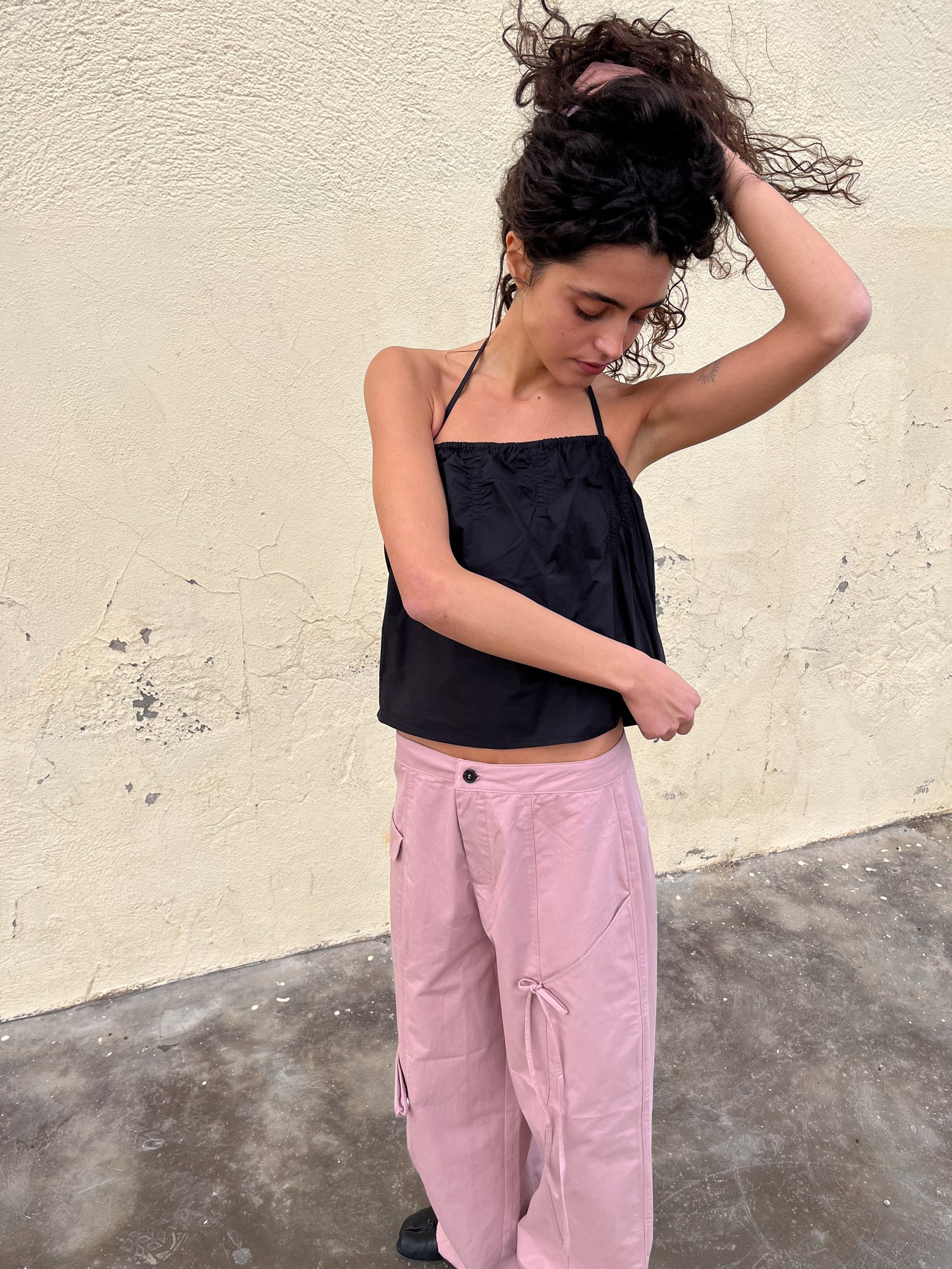 A woman stands against a textured beige wall, wearing the black Tube Top and loose, light pink pants. She looks down as she lifts her curly hair with one hand, while her other arm crosses her body.