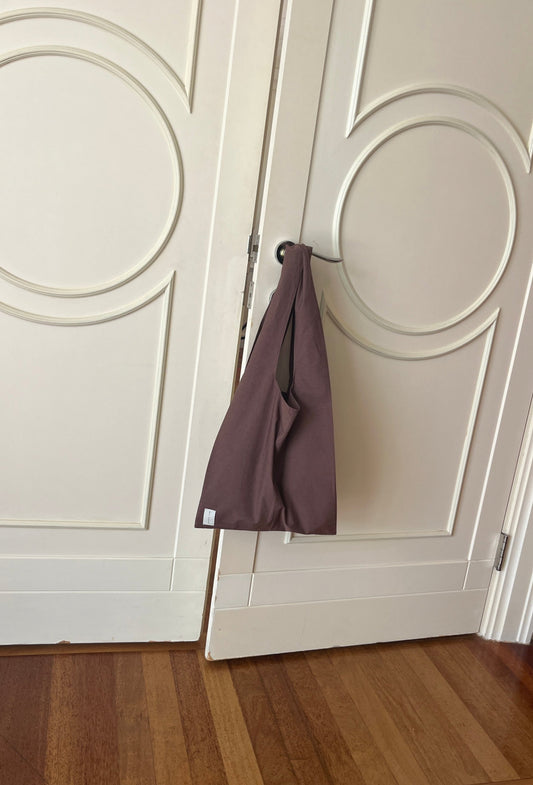 A Seam Tote in mauve hangs from a silver handle on white double doors with circular patterns, above a wooden floor.