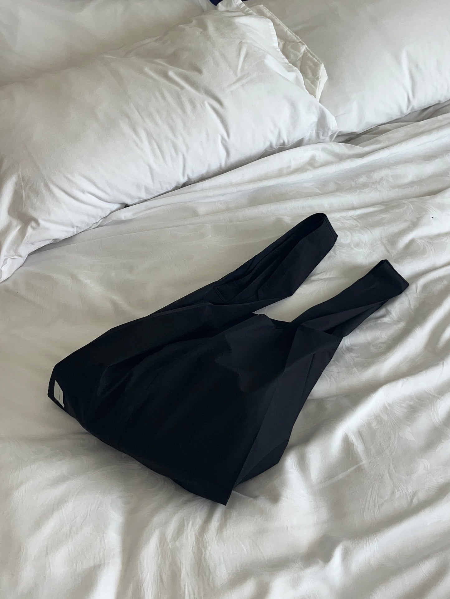 The Seam Tote - Black by Deiji Studios, crafted from organic cotton for everyday ease, features two handles and rests on a white bed with white pillows in the background.