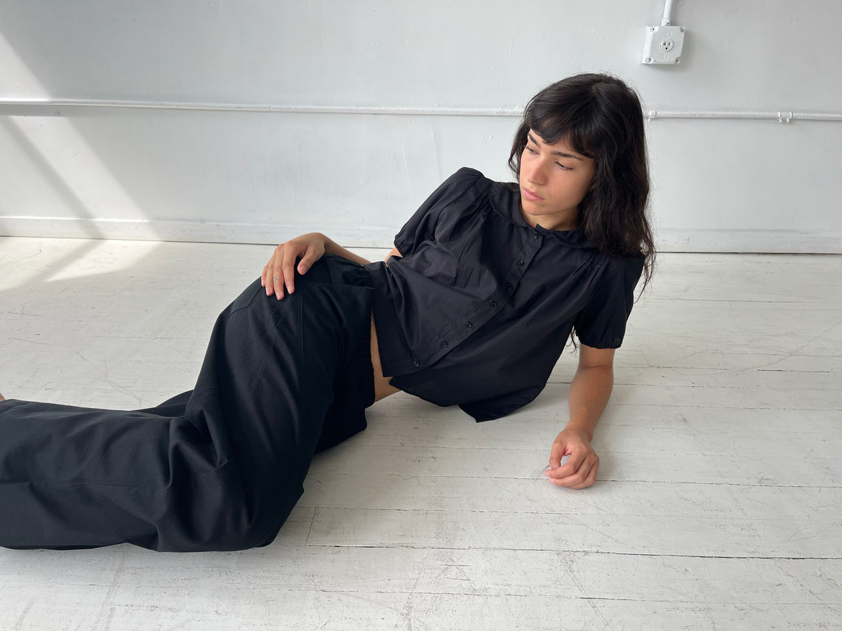 A woman reclines on a white studio floor, wearing the Petal Top - Black by Deiji Studios, a cotton voile top with a peter pan collar and puffed short sleeves, styled with the Curve Pant in Black.