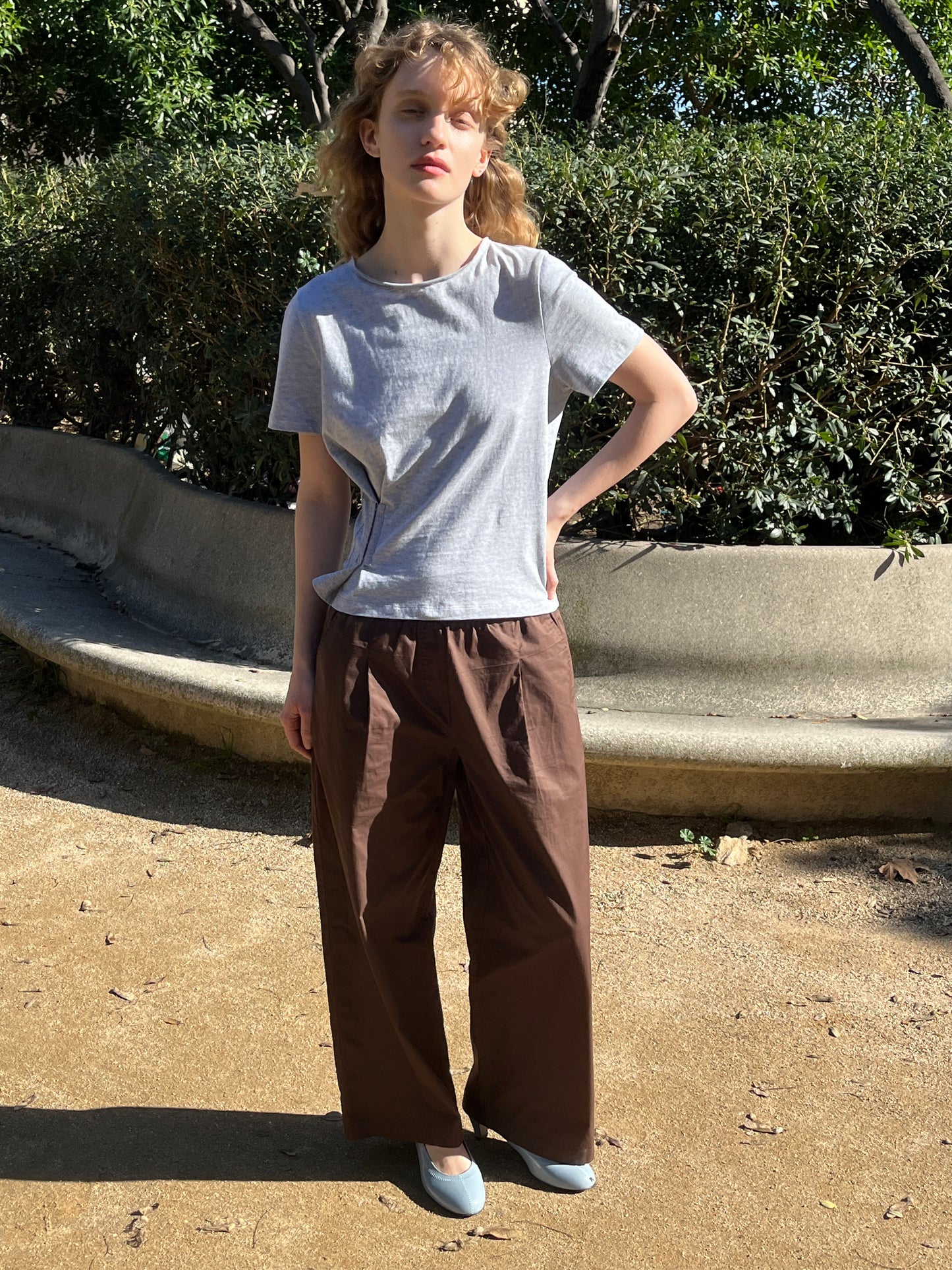 A person with curly blonde hair stands outdoors on a sunny day, wearing a light gray t-shirt and Deiji Studios Contrast Waist Pant - Brown with a relaxed straight leg, light blue shoes, one hand on their hip, and trees and bushes in the background.
