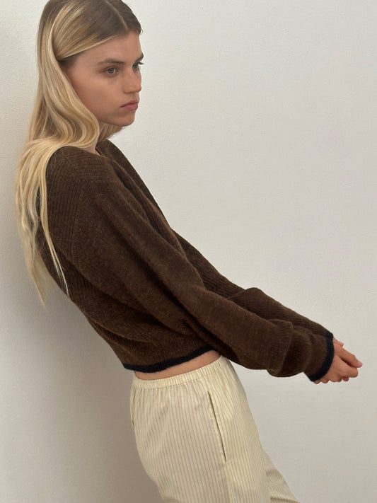 A young woman with long blonde hair wears the Cardi - Tawny, standing sideways against a plain off-white wall and looking slightly downward with a neutral expression.