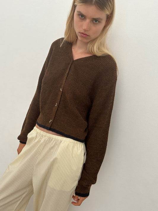 A woman with long blonde hair wears the Cardi - Tawny by Deiji Studios and loose, cream-striped pants, standing against a plain white background and looking slightly down.