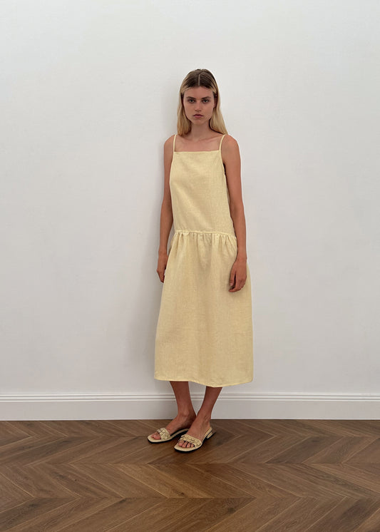 A woman with straight light brown hair and a neutral expression stands before a white wall on a herringbone wooden floor, wearing the Gathered Halter Dress in Butter Yellow with adjustable tie straps and flat sandals.