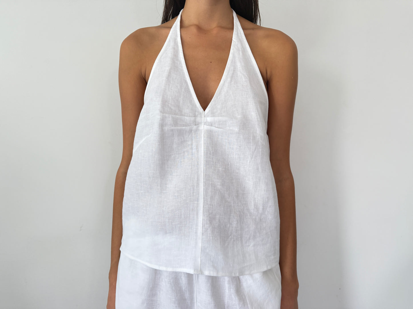 A person wearing the Linen Pinched Halter Top - White, a sleeveless French linen halter with a V neckline, stands against a plain white background. Only the upper body is visible.