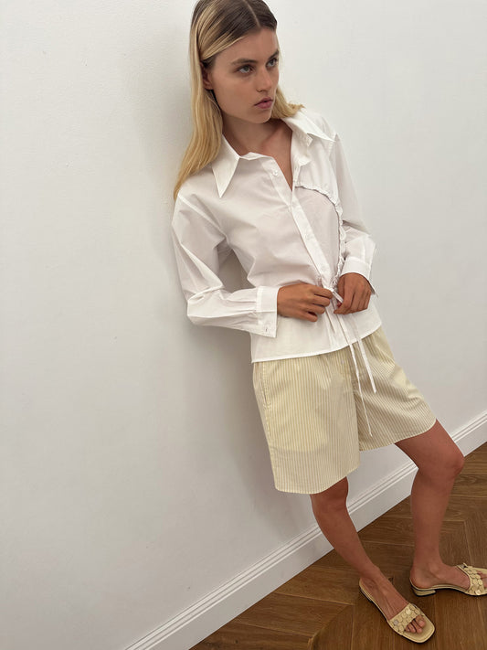 A woman with long blonde hair stands indoors against a white wall, wearing the Deiji Studios Arch Tie Shirt in white and light beige pinstriped shorts, paired with light-colored open-toe sandals on a wooden floor.