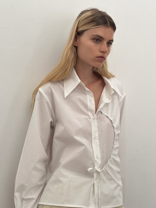 A young woman with long blonde hair wears the Deiji Studios Arch Tie Shirt in white, made from organic cotton with an asymmetrical curved seam. She stands against a plain white wall, gazing to the side with a neutral expression.