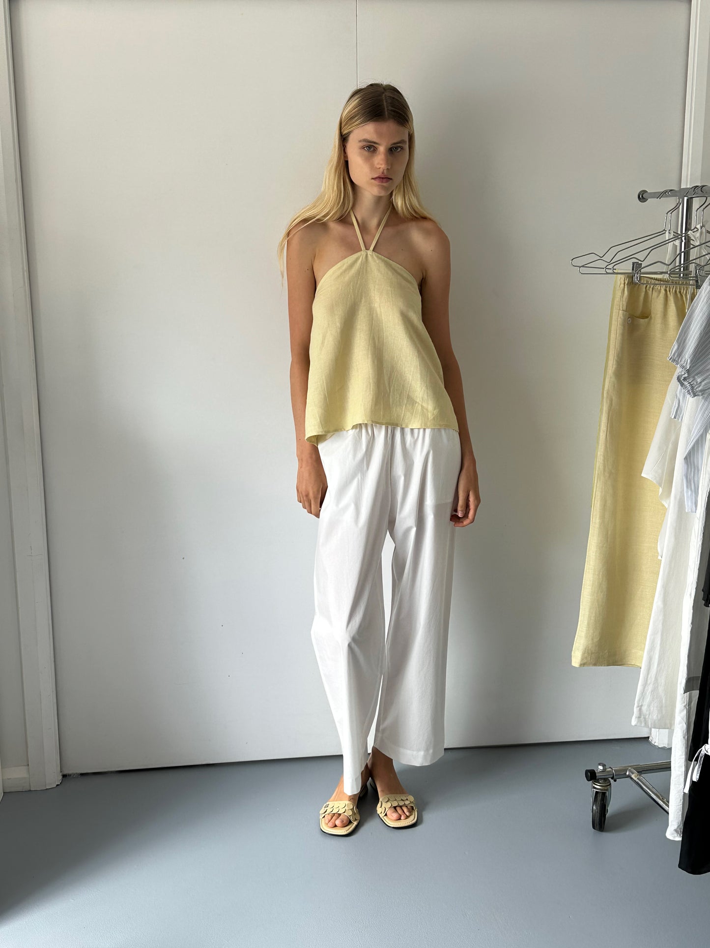 A woman stands indoors by a white wall, wearing a light yellow halter top, Deiji Studios Ease Trouser - White in organic cotton, and sandals. A clothing rack with light garments is nearby.