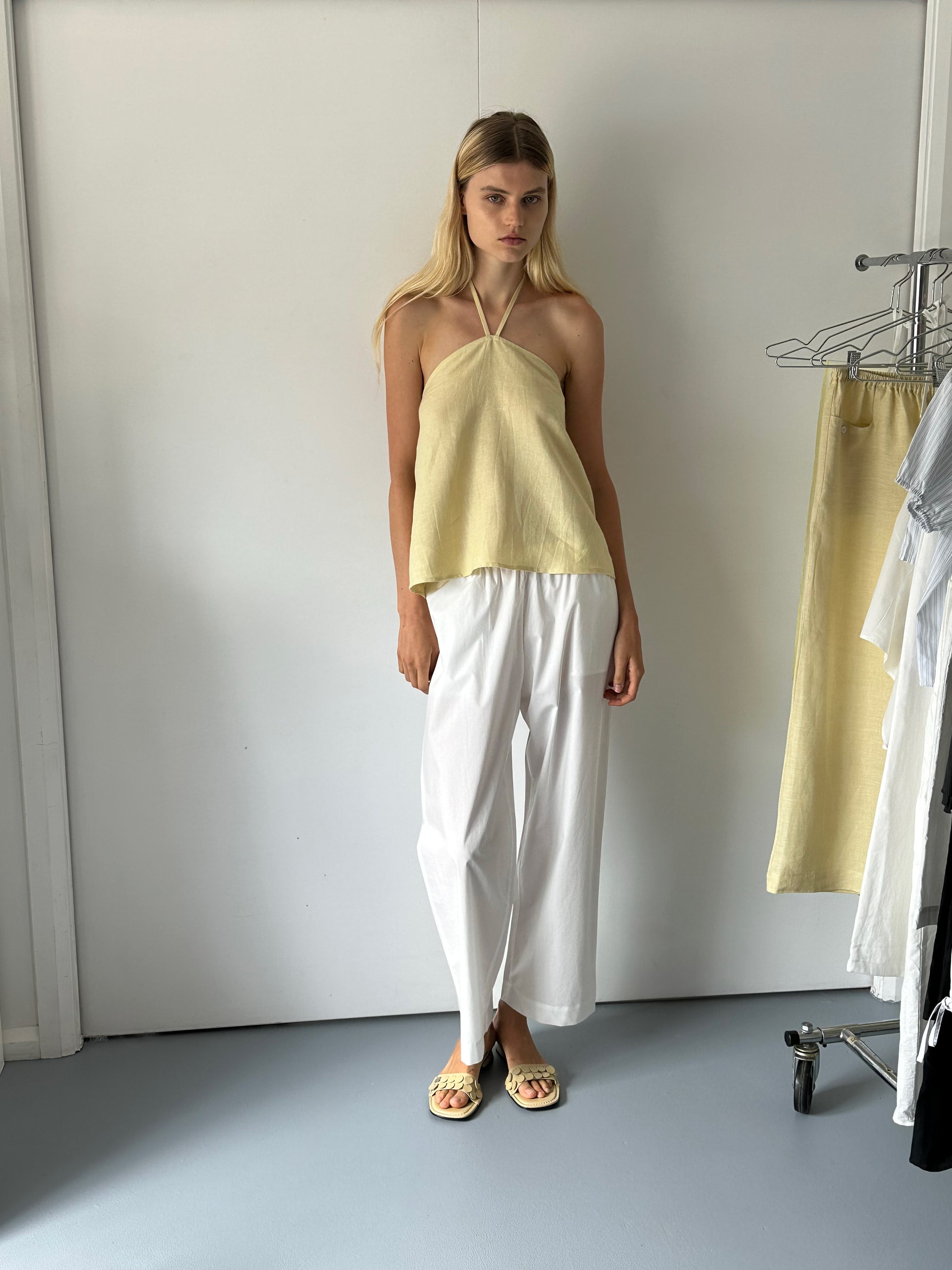 A woman stands indoors by a white wall, wearing a light yellow halter top, Deiji Studios Ease Trouser - White in organic cotton, and sandals. A clothing rack with light garments is nearby.