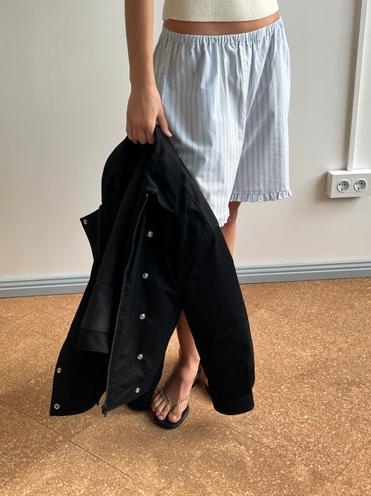 Wearing a white tank top, light blue pinstriped shorts, and flip-flops, a person stands indoors holding the Lapel Jacket - Black by Deiji Studios; only their lower half is visible on a brown floor near a white wall.