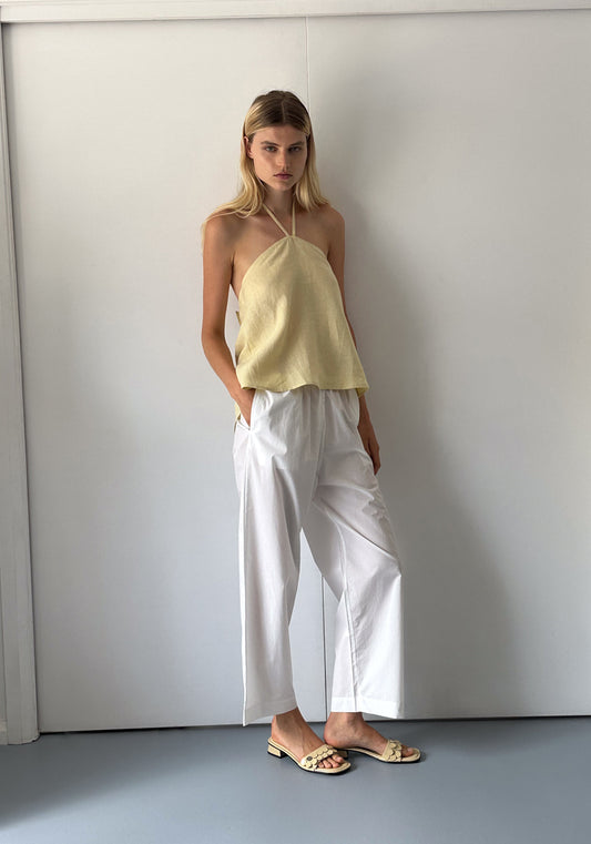 A woman with straight blonde hair stands indoors by a white wall, wearing the Linen Wrap Back Top - Butter Yellow by Deiji Studios, loose white pants, and flat sandals. Her hands are in her pockets and she has a neutral expression.