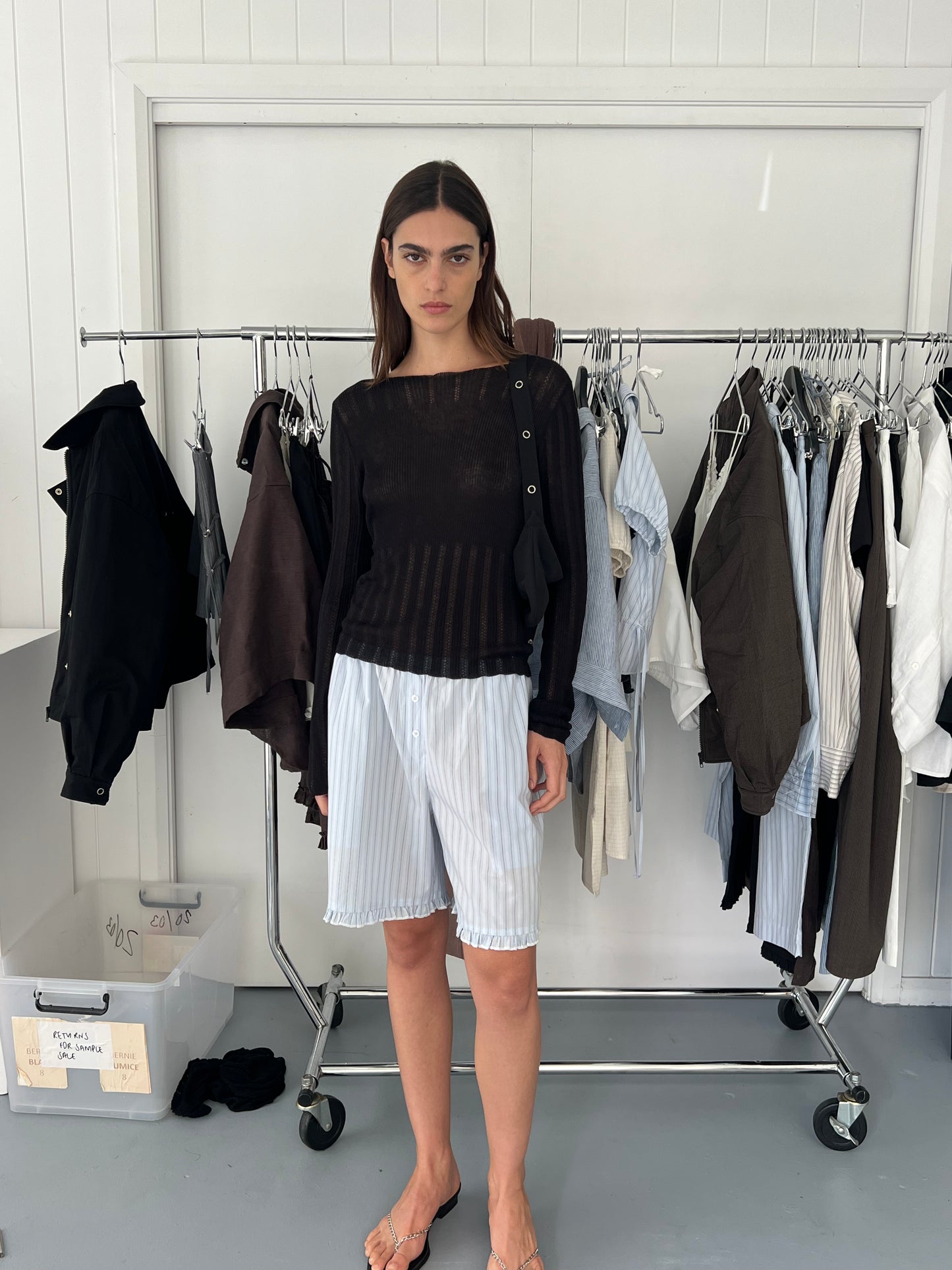 A woman stands indoors before a clothing rack, wearing the Deiji Studios Open Work Longsleeve in black and loose, knee-length white striped shorts. Various garments hang behind her, with a box on the floor to her left.