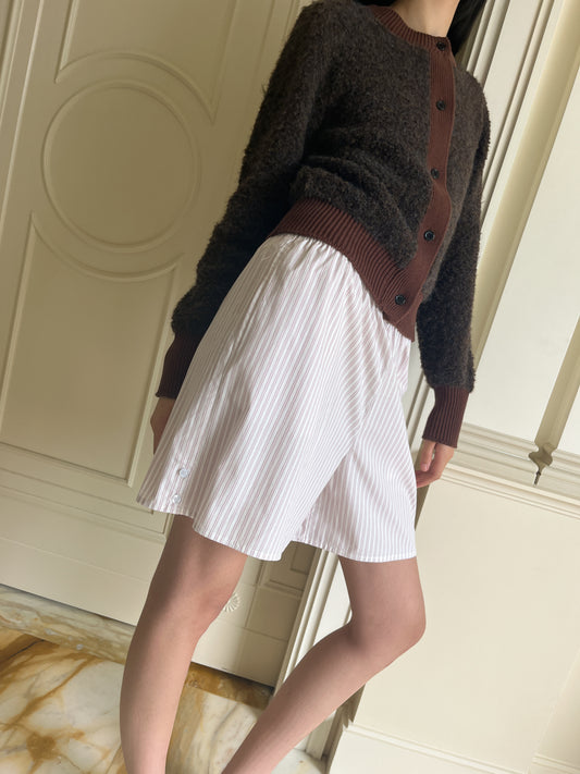 Someone models the Button Short, a dark brown, buttoned cardigan paired with a white knee-length striped skirt. They stand indoors by a cream-paneled wall and marble floor. The photo does not show their head.