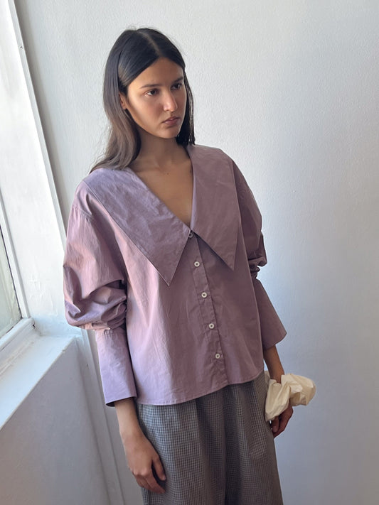 A woman with long dark hair stands indoors by a window, wearing an Oversized Collared Shirt - Mauve with baggy sleeves and gray checkered pants. She holds a white cloth in her left hand.