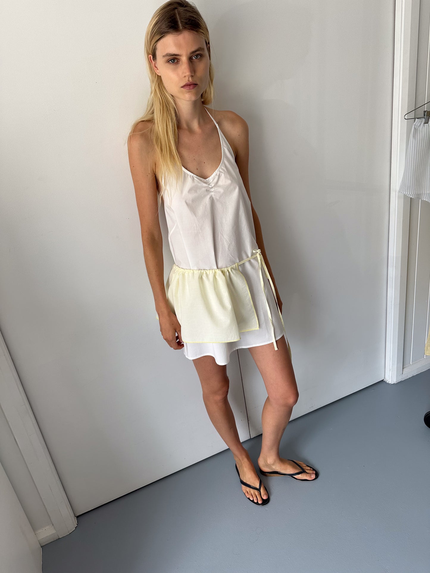A woman with long blonde hair stands indoors against a white wall, wearing the 0 Round Halter Dress in white with a light yellow sash and black flip-flops. She has a neutral expression and relaxed posture.