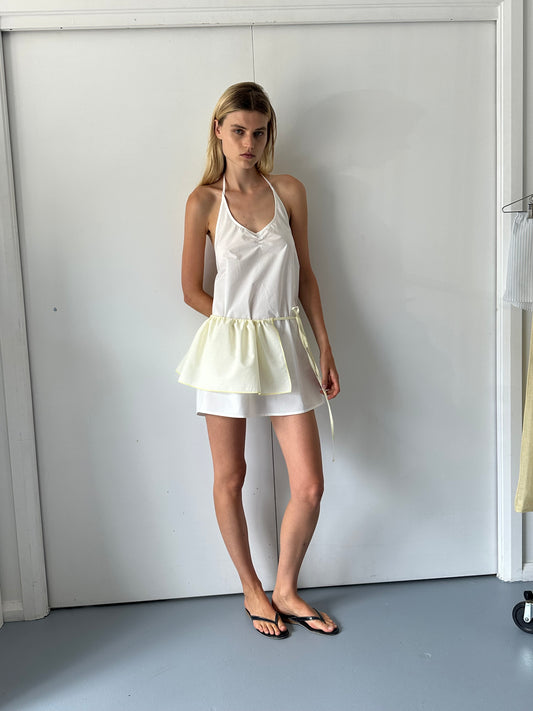 A woman with straight blonde hair stands indoors against a white wall, wearing a sleeveless white dress, the Peplum Belt - Lemon at her waist, and black flip-flops. She has a neutral expression.