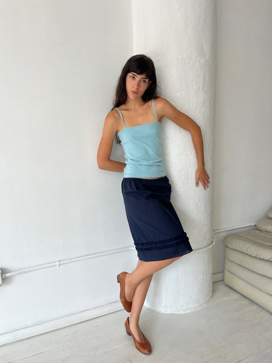 A woman leans against a white column, wearing the Daytime Skirt - Navy by Deiji Studios, a cotton voile midi skirt with double frill detailing at the hem, styled with the Scallop Tank in Sky and brown flats.