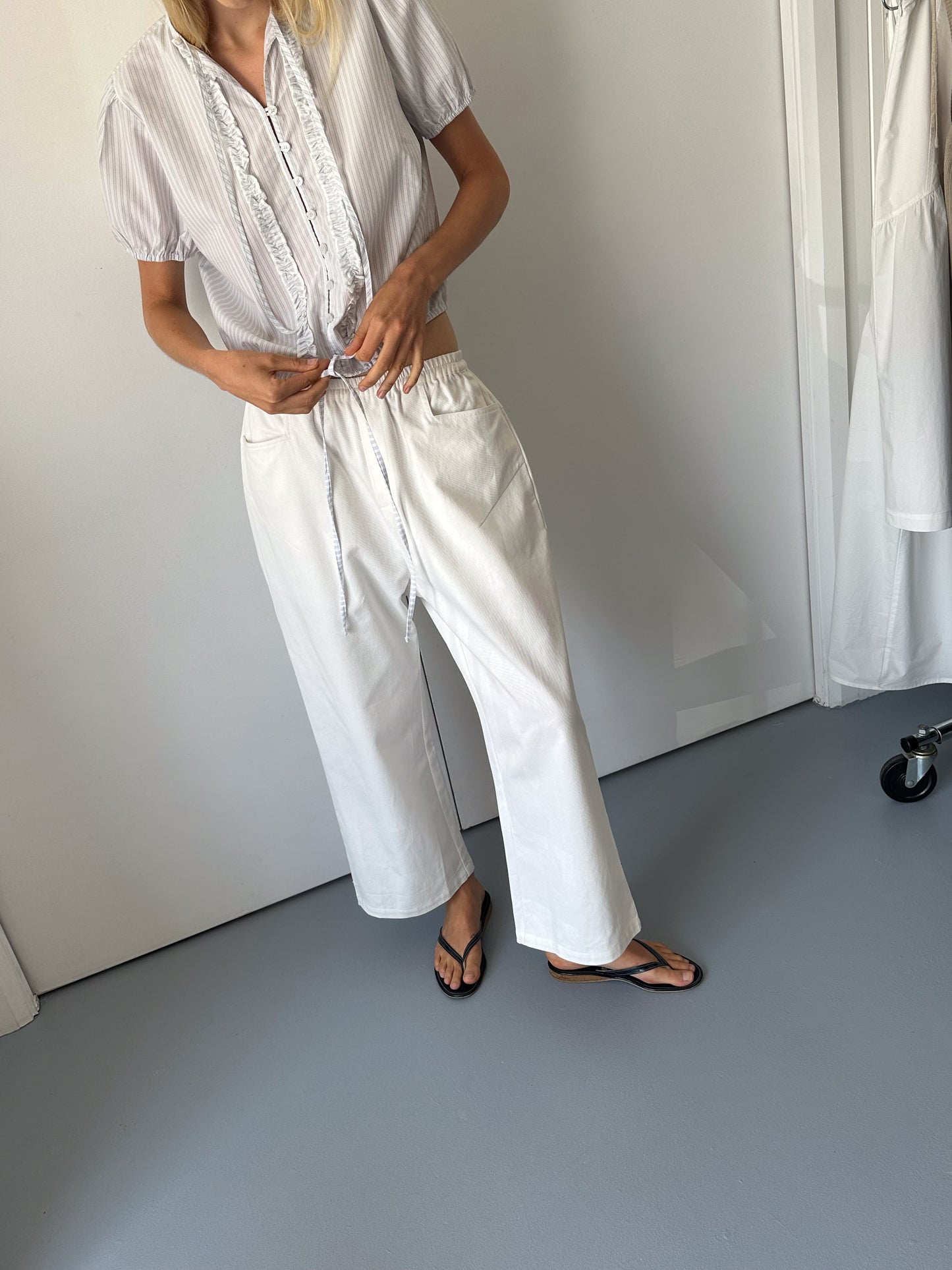 A person stands indoors on a gray floor, adjusting the drawstring ties of a short-sleeved, light-striped blouse. Theyre wearing 0s Square Pocket Pant in white with black flip-flops. Their face is out of frame.