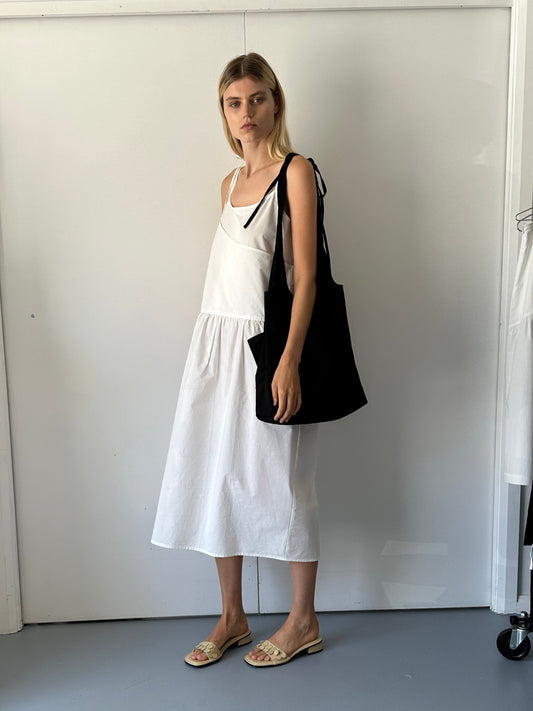 A woman with straight blonde hair and a neutral expression poses indoors against a white background, wearing the Double Layer Dress - White, an organic cotton sleeveless midi with adjustable straps, paired with tan sandals and a large black shoulder bag.