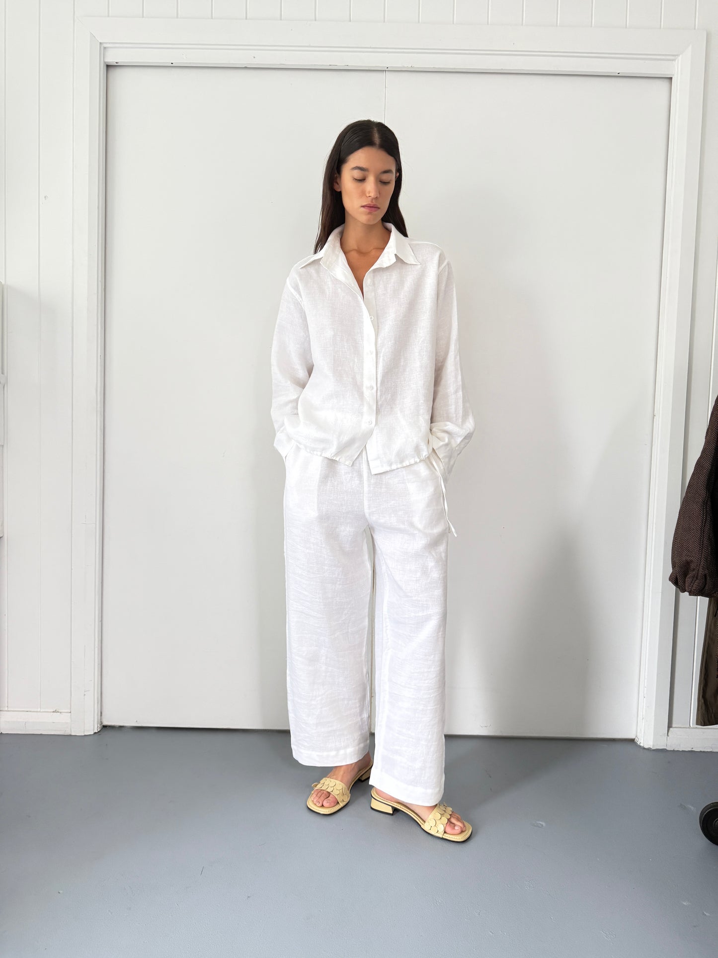 A woman with long dark hair stands indoors by a white door, wearing the Tie Split Set - White: a relaxed-fit organic linen shirt with matching wide-leg pants, paired with light-colored sandals.