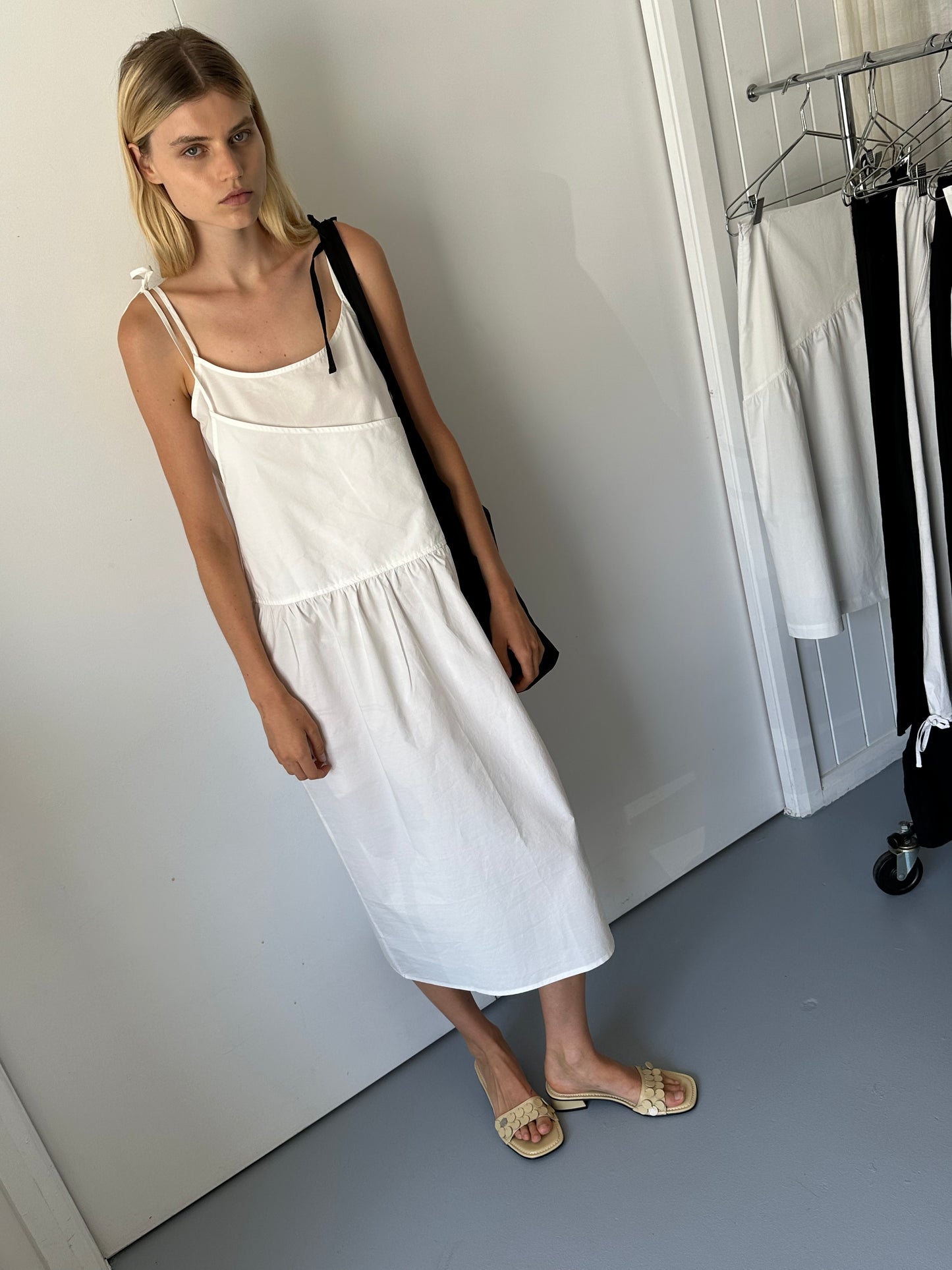A woman with straight blonde hair stands indoors wearing the Double Layer Dress - White and beige sandals, carrying a black bag, near a wall with hanging clothes and a clothing rack.