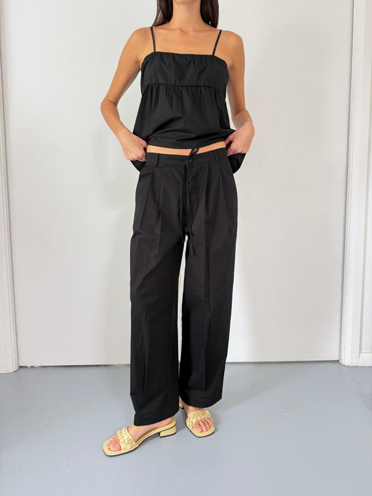 A woman stands indoors against a plain white background wearing Deiji Studios’ Turn Pant in Black with an adjustable drawcord, paired with a black spaghetti strap top and light yellow open-toed sandals, her hands in the pockets.