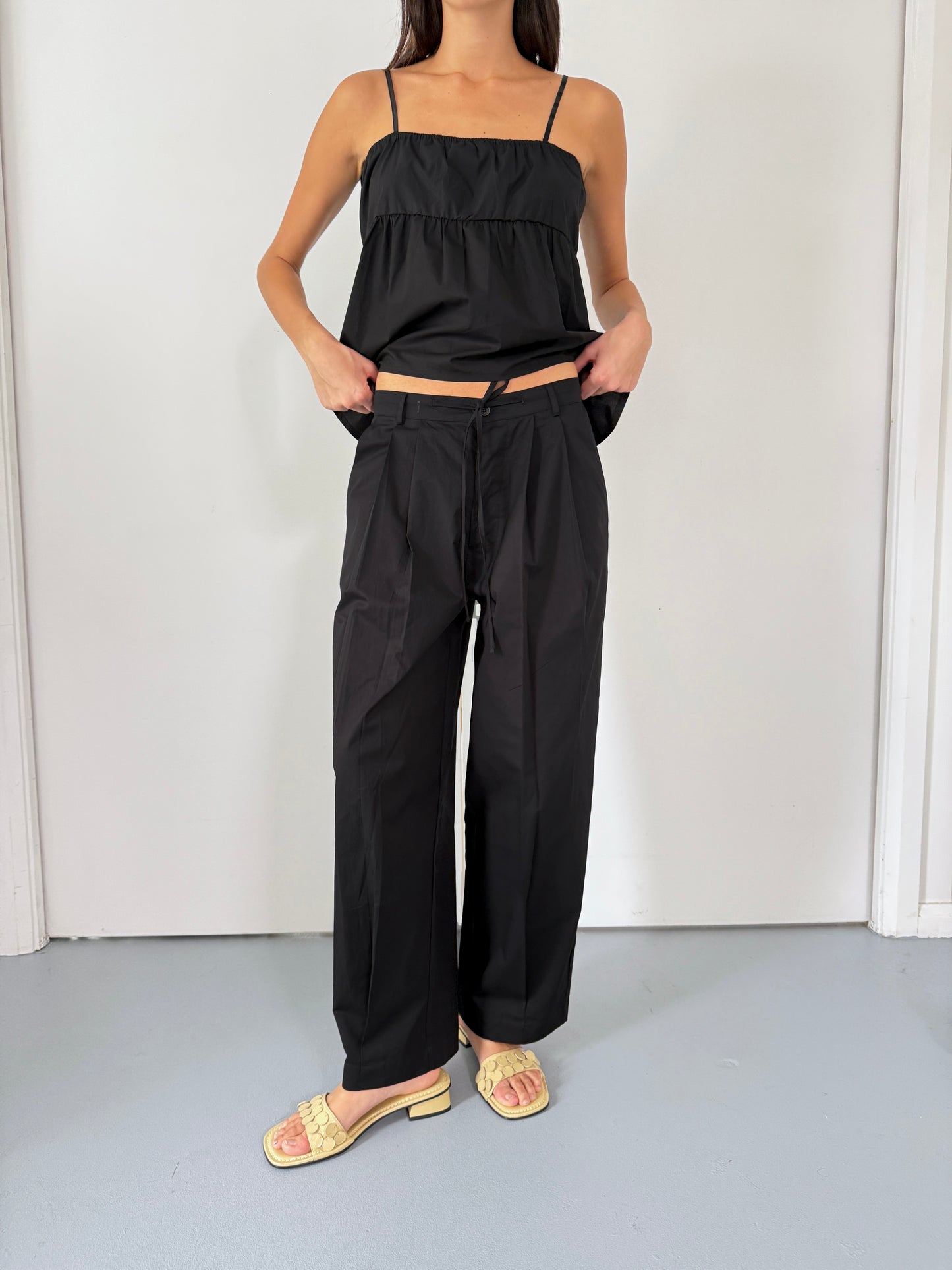 A woman stands indoors against a plain white background wearing Deiji Studios’ Turn Pant in Black with an adjustable drawcord, paired with a black spaghetti strap top and light yellow open-toed sandals, her hands in the pockets.