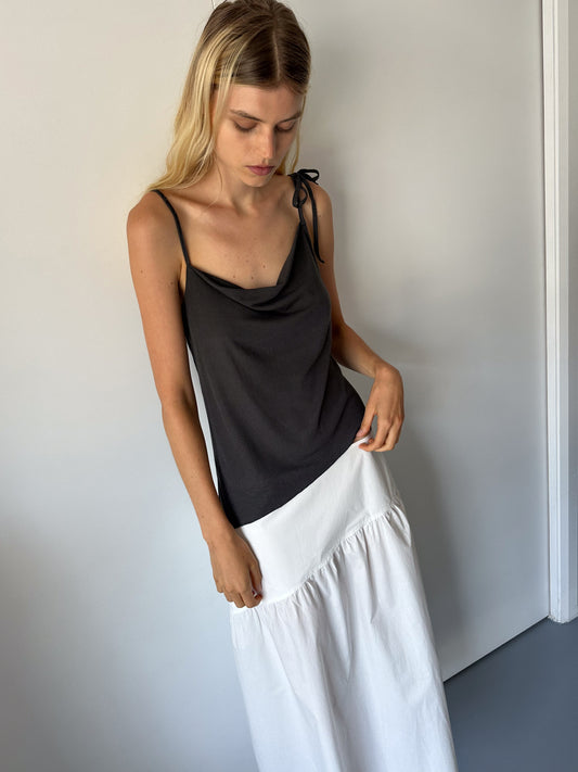 A woman with long blonde hair wears the Deiji Studios Split Top in Night Blue with a shoulder tie and a high-waisted white skirt. She stands against a plain light wall, looking down as she lightly holds her skirt.