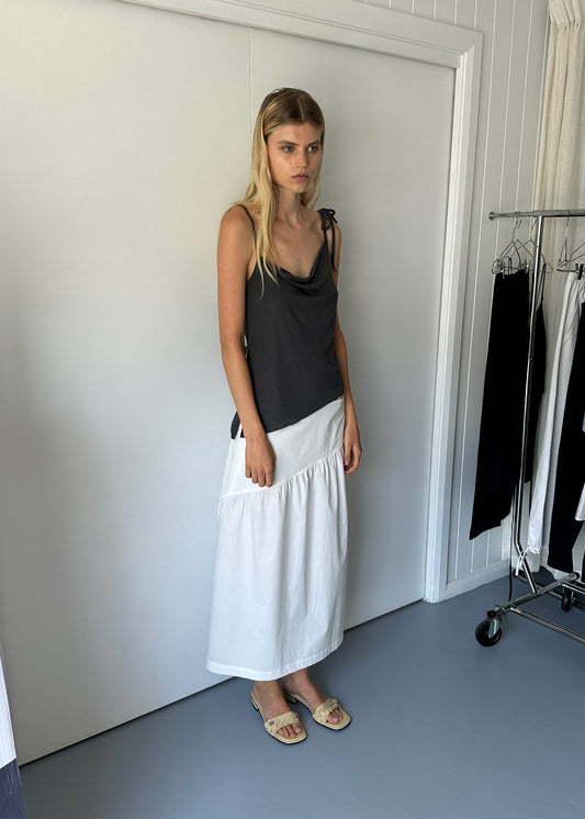 A woman with long blonde hair stands indoors wearing a Deiji Studios Split Top - Night Blue and a white maxi skirt beside a clothing rack with black and white garments. The room features light walls and a gray floor.