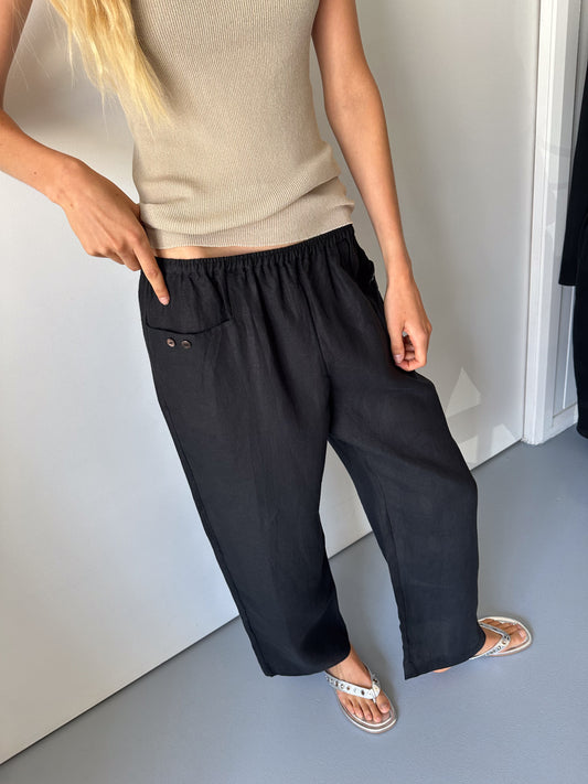 A person wearing a beige sleeveless top, flip-flops, and Deiji Studios' Linen Square Pocket Pant in black points to the pant's pocket button. The background is minimal with white and gray walls.