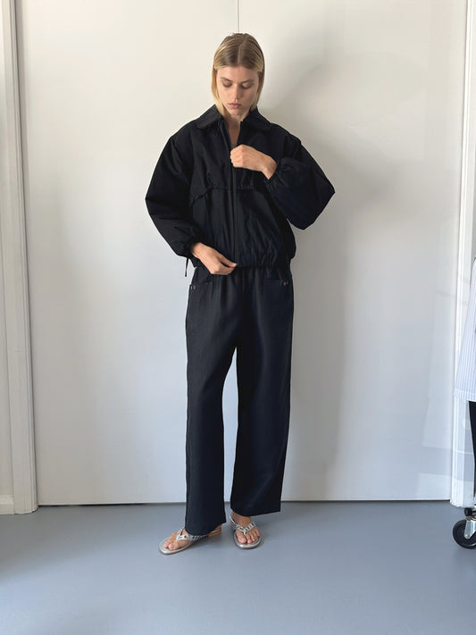 A person stands indoors against a white wall, wearing a loose black jacket and Deiji Studios' Linen Square Pocket Pant in organic French linen with silver sandals. They look down, holding the jacket collar with one hand.